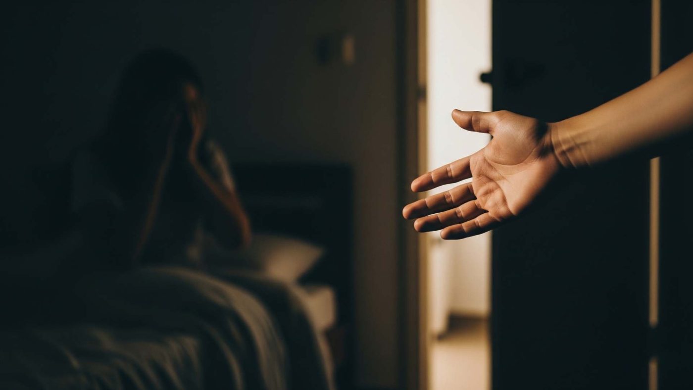 Loş bir odada, yatağında korku içinde doğrulmuş (ama yüzü net görünmeyen, daha çok bir silüet gibi) bir çocuk veya yetişkin. Odanın kapısının aralığından sızan hafif bir ışık huzmesi, bu silüetin üzerine düşer. Ön planda, net bir şekilde görünen, sakin ve şefkatli bir elin (ebeveyn veya partnerin eli) yatağın kenarına uzandığı bir kompozisyon. Bu görsel, "Yalnız değilsin, güvendesin" mesajını doğrudan ve güçlü bir şekilde verir. Odak noktası korku değil, varlığıyla sakinlik getiren "koruyucu el"dir.